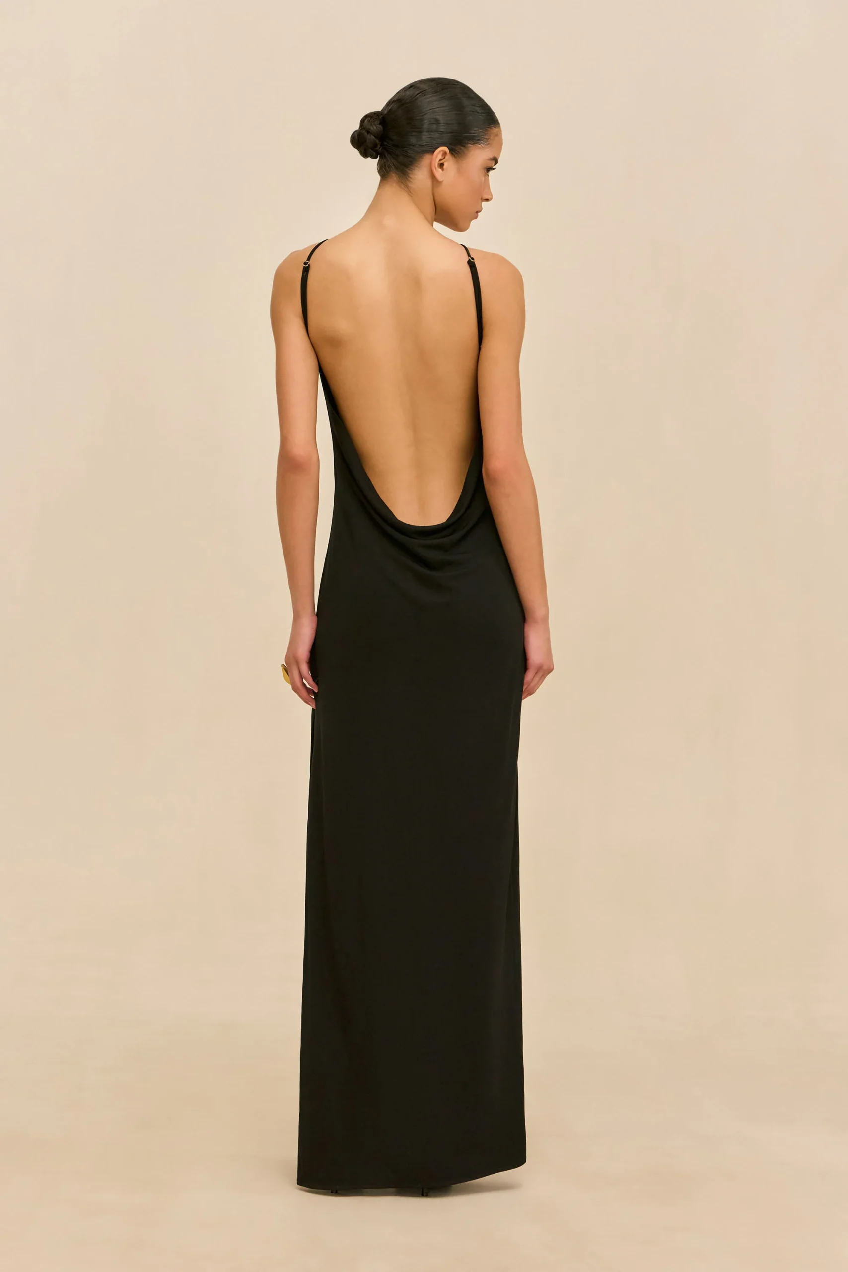 IMG_1537 - New Trend Woman in a long black backless dress, viewed from behind, with a neat bun in her hair against a beige backdrop.