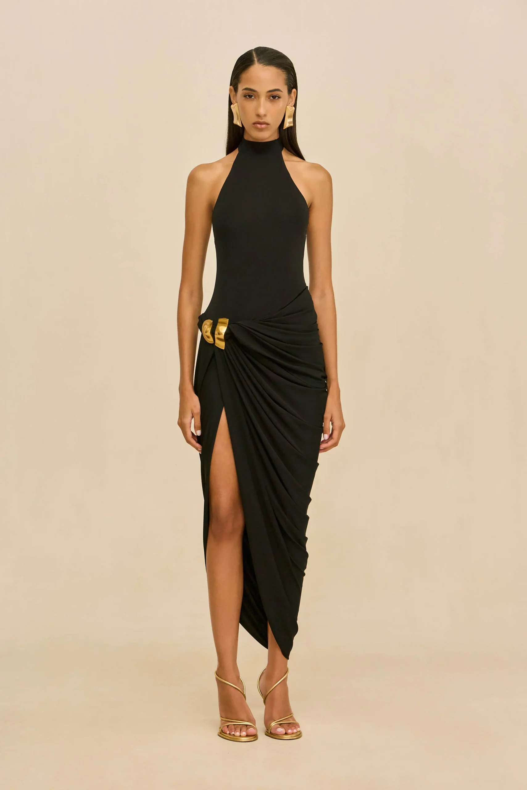 IMG_1560 - New Trend Female model wearing a black halter-neck gown with a draped skirt and gold waist accents, standing against a beige background.