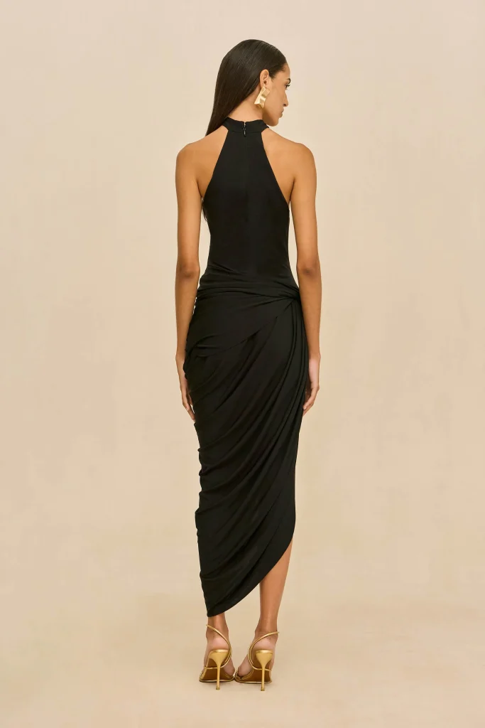 Woman in a black halter dress viewed from behind, draped skirt, gold earrings, and strappy gold heels against a beige backdrop.