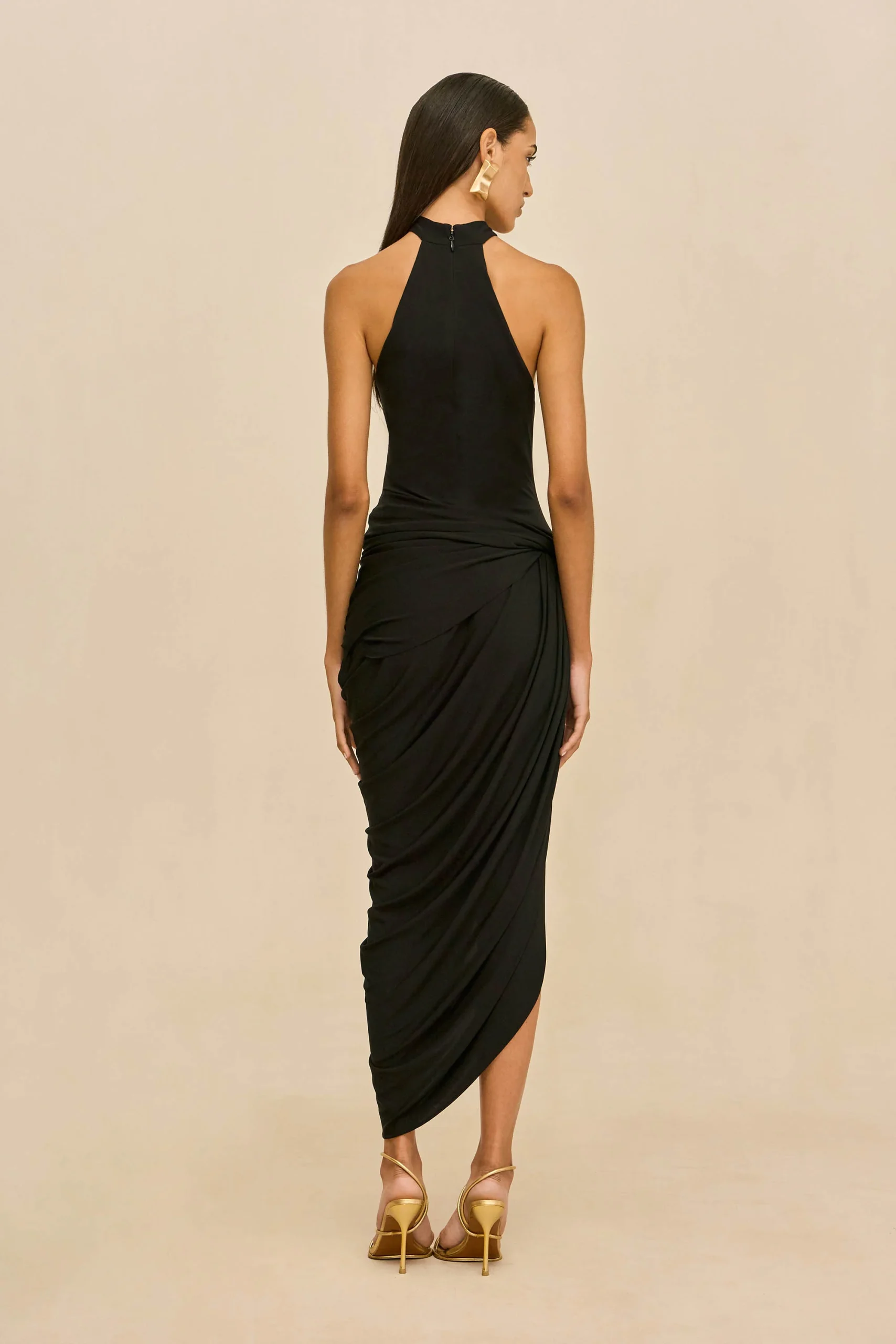 IMG_1564 - New Trend Woman in a black halter dress viewed from behind, draped skirt, gold earrings, and strappy gold heels against a beige backdrop.