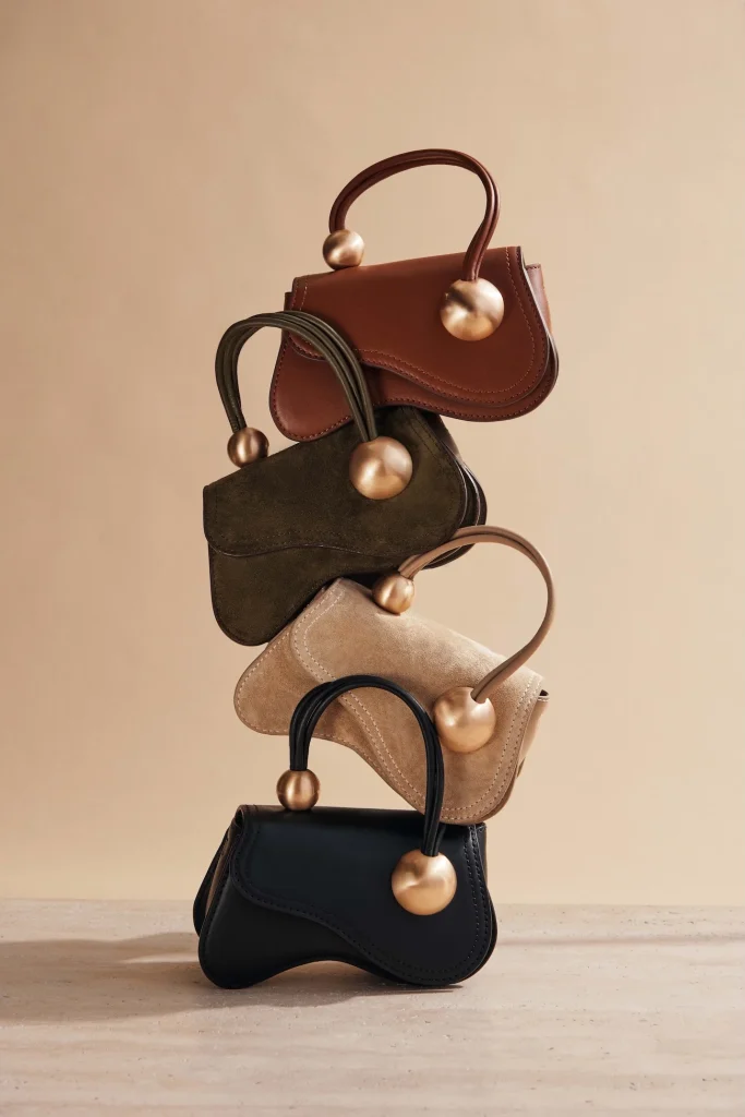 Stacked small handbags in brown, olive, beige, and black with curved handles and gold spherical hardware on a neutral backdrop (fashion product display).