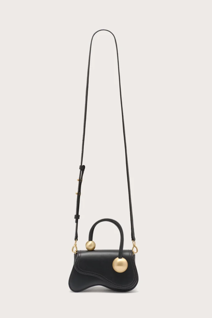 Black leather handbag with curved bottom and gold ball charms on the strap attachments, hanging from a long strap