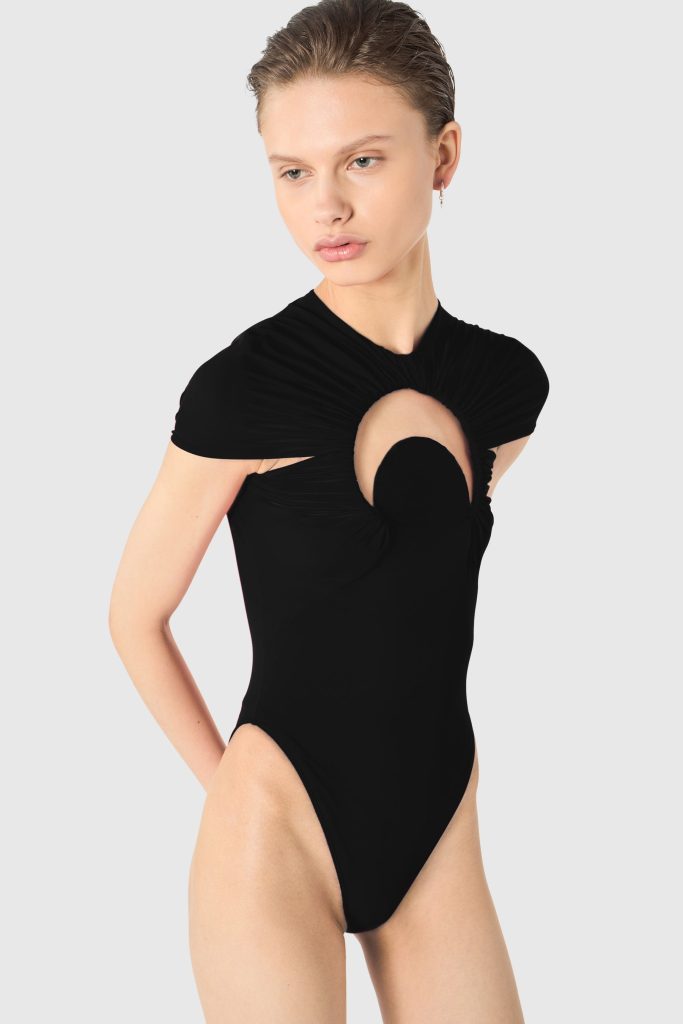 Model wearing a black one-piece swimsuit with a circular chest cut-out, posing against a light gray background