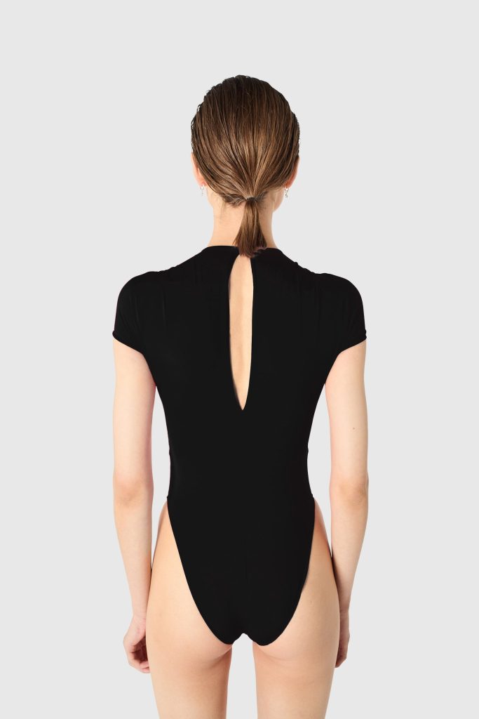 Back view of a woman in a black one-piece swimsuit with a vertical back cutout and hair tied in a low ponytail