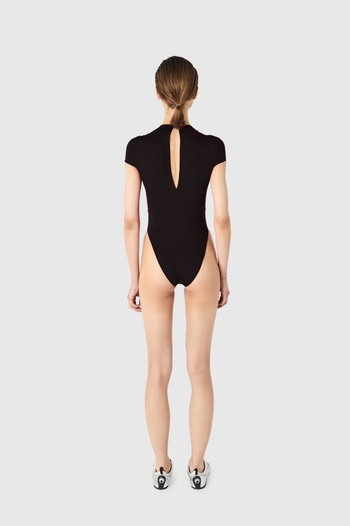 Back view of a woman wearing a black one-piece bodysuit with a teardrop back opening, her hair in a low ponytail, white sneakers visible.