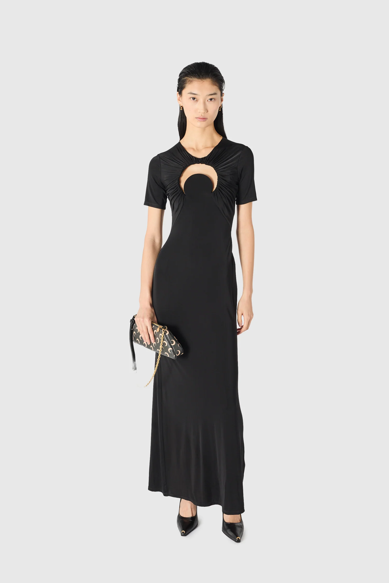 IMG_1621 - New Trend Model wearing a black floor-length dress with a circular cutout at the chest, holding a patterned clutch.
