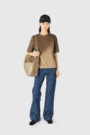 Woman standing against a light gray background, wearing a brown short-sleeve top, blue patterned wide-leg jeans, a black cap, and carrying a beige woven tote.