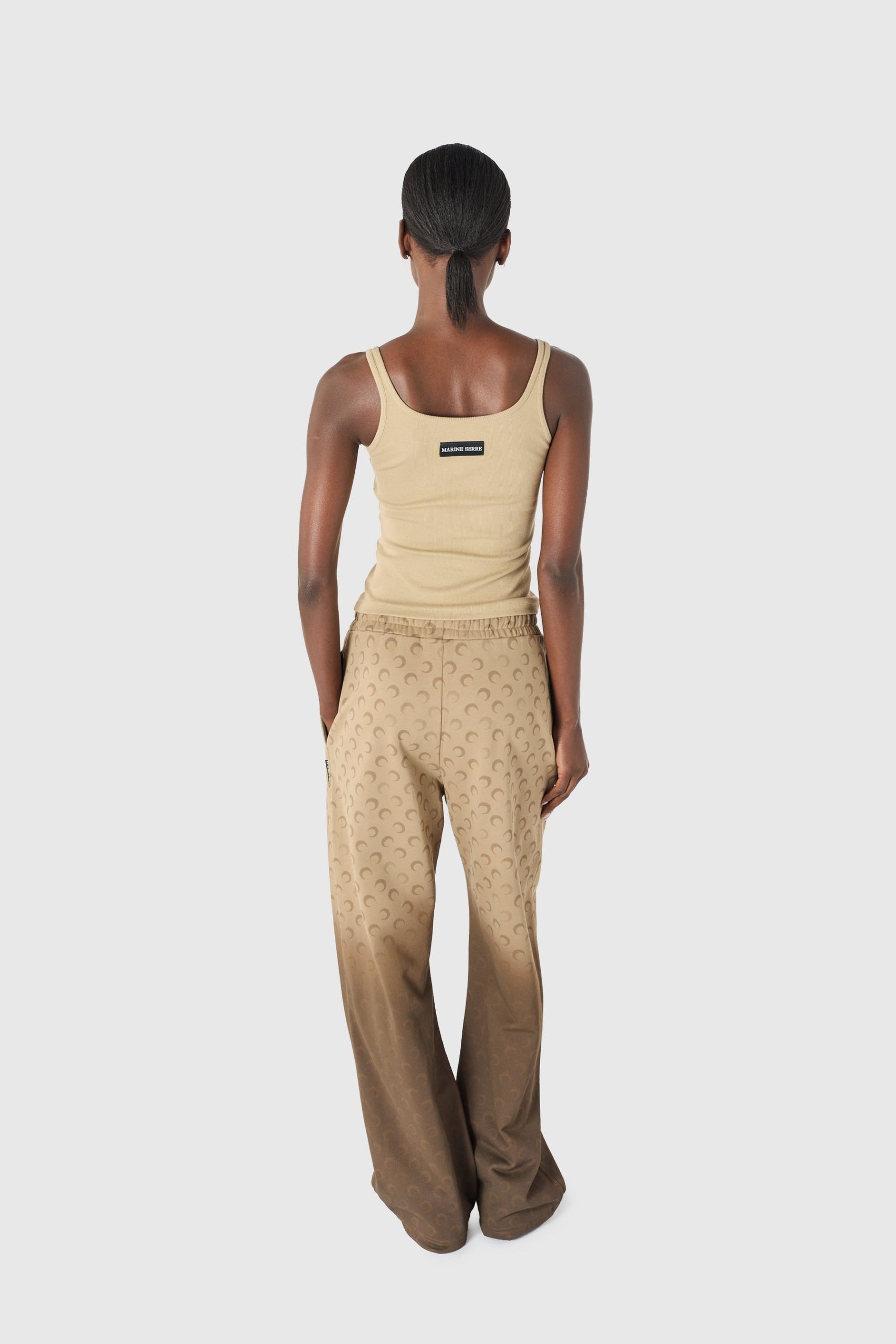 IMG_1640 - New Trend Back view of a person wearing a beige tank top and tan patterned pants, with a small label on the tank top's back and hair tied in a low ponytail.