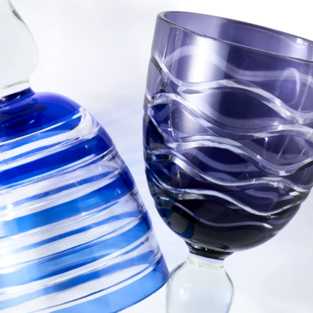 Close-up of two curved glass bottles, one blue with white stripes and one purple-tinted, against a pale background.