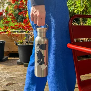 Person in a blue outfit holds a silver stainless steel water bottle with a cartoon girl illustration outdoors on a wooden deck, red chair nearby.