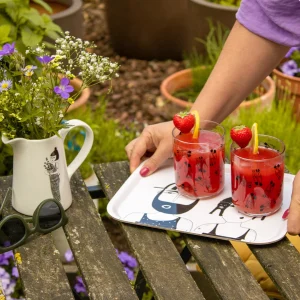 Two strawberry cocktails with lemon wedges on a illustrated tray in a garden, a hand reaching toward the drinks beside a vase of flowers and sunglasses on a weathered wooden table.