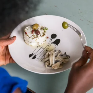 A plate holds a small round herb-crusted dessert topped with olives and flowers, with mushroom pasta on the side and a fork picking up a green olive.