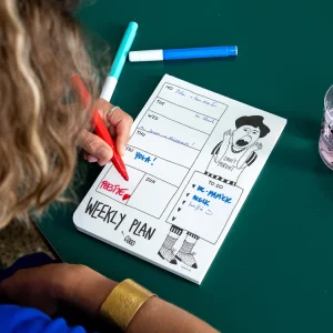 Weekly planner open on a green table with doodles, as a person writes with a red marker.