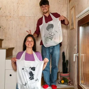 Smiling couple wearing white aprons with cartoon drawings, posing on indoor stairs.