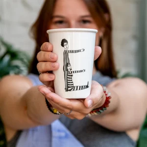 Close-up of a white cup held toward the camera, featuring a black line-drawing of a tall and a shorter figure in striped outfits with arms outstretched.