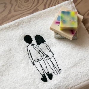 White towel with a black-embroidery design of two naked women standing side by side; colorful soap bars rest in the top-right corner.