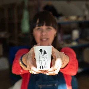 Woman holding a white cup toward the camera, cup printed with a simple line drawing of two people holding hands.