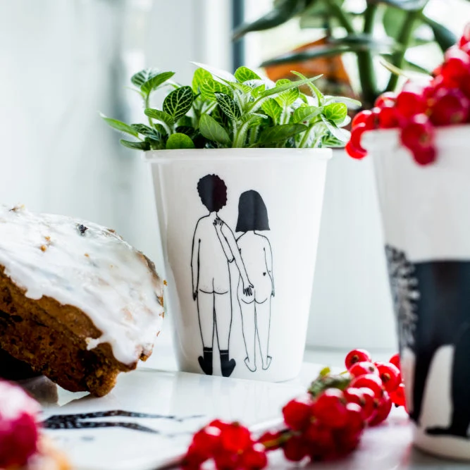 White cup with a black illustration of a couple holding hands, filled with a green plant, on a kitchen counter near cake and red berries.