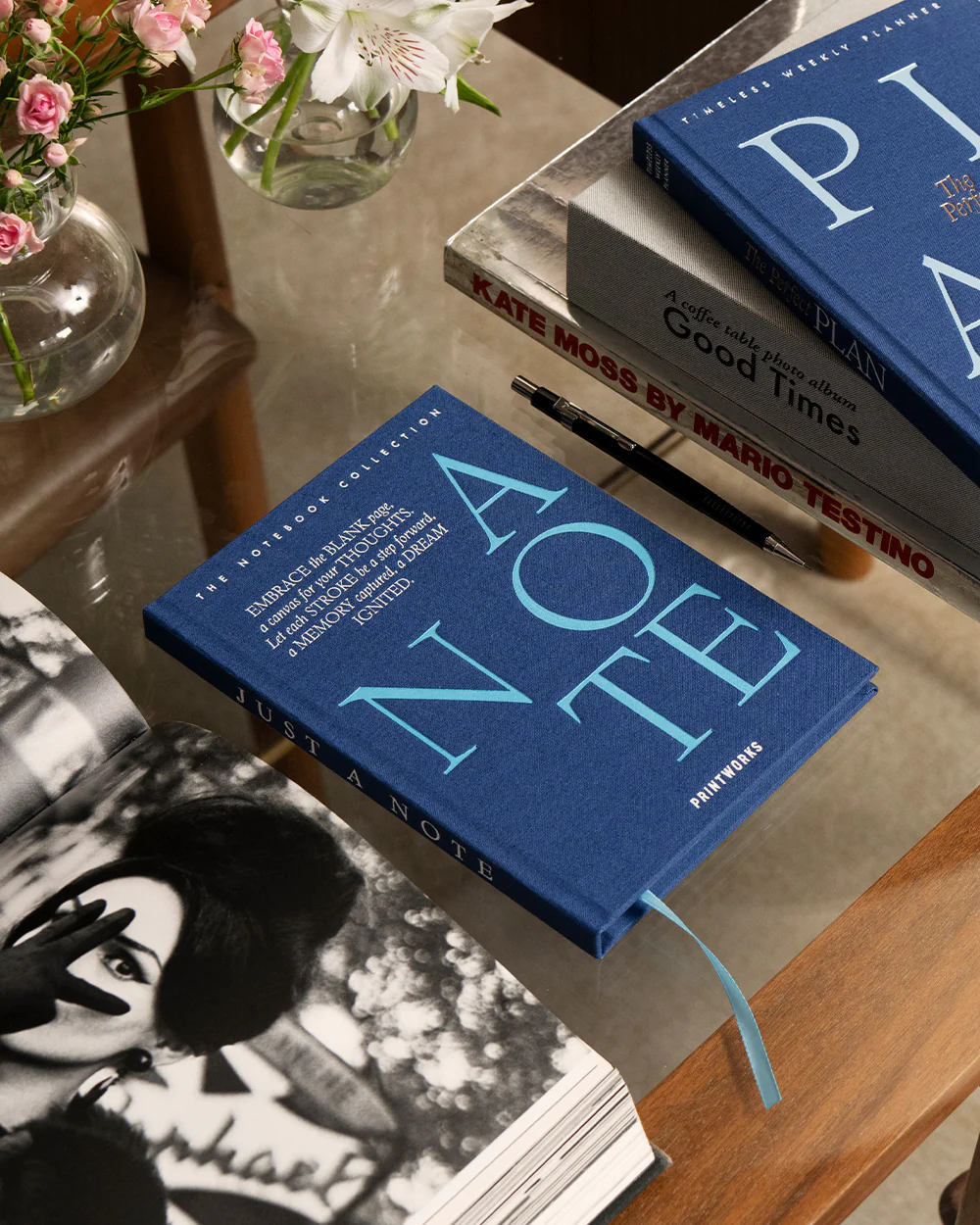 IMG_2410 - New Trend Blue hardcover notebook titled 'A NOTE' on a glass table, beside a stack of photography books (e.g., Kate Moss by Mario Testino) and a black pen; pink flowers in vases visible in the background.
