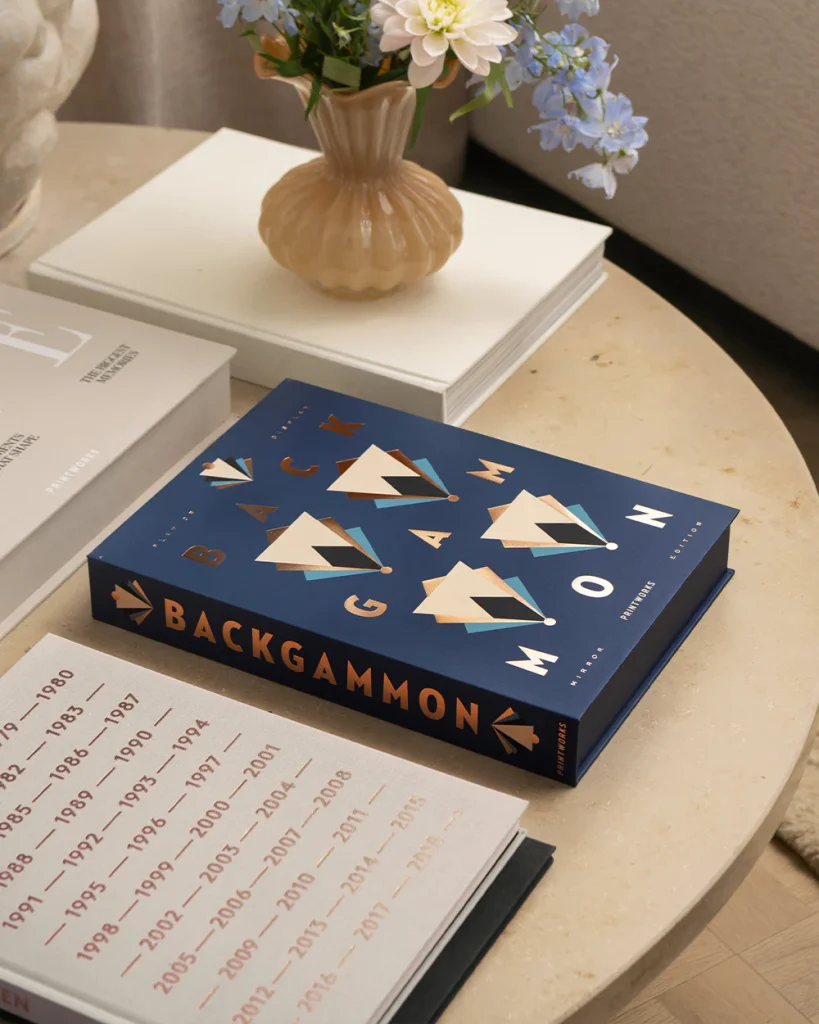 Blue Backgammon book with geometric shapes on a marble table, beside a vase of flowers and white stacked books, in a cozy setting.
