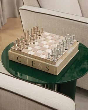 Chessboard with beige and white squares, set on a large beige book titled CHESS on a round green glass table near beige seating.