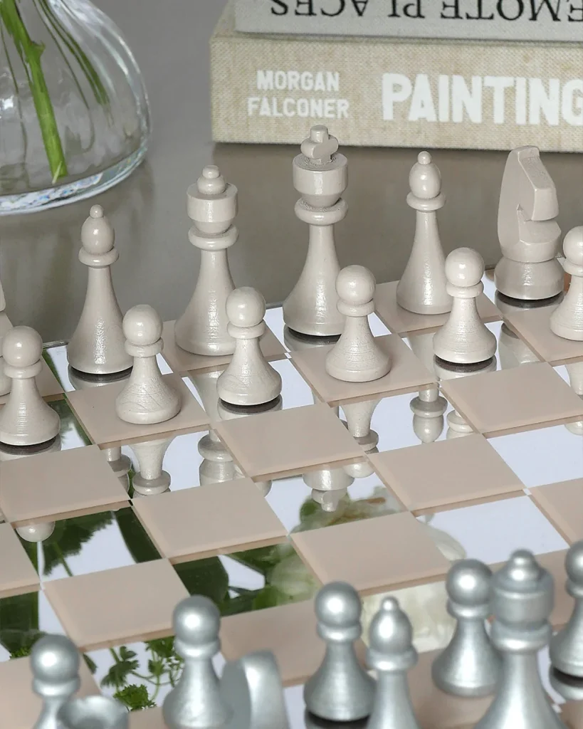 Close-up of a chessboard with white pieces on a mirrored surface; reflections multiply the pieces, with stacked books in the background showing 'PAINTING'.