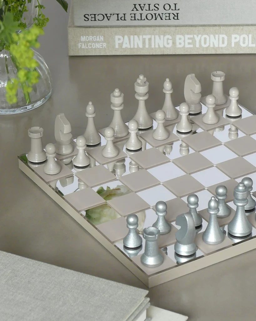 A chessboard with white and silver pieces set up on a glossy surface, with a vase of greenery and stacked books in the background.