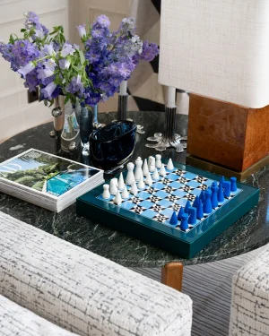 Blue and white chessboard set up for a game on a dark marble table, with white and blue pieces arranged opposite each other. Some purple flowers in a vase and a magazine sit nearby.
