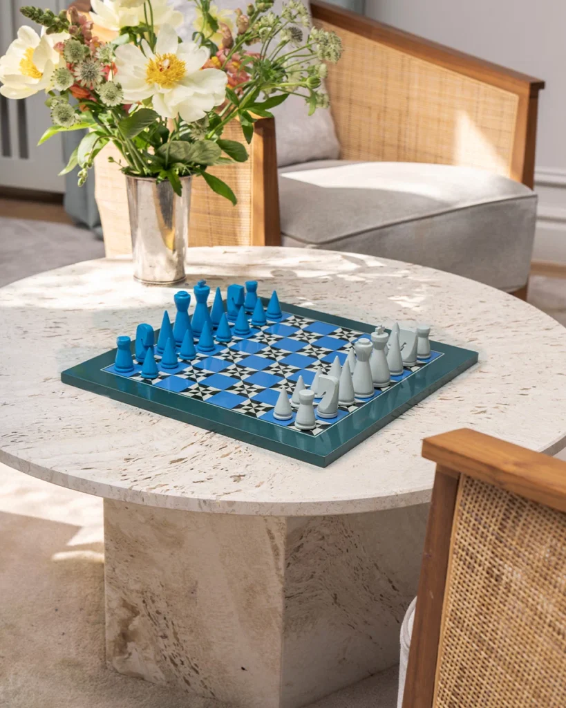 Blue chess pieces on a patterned board set on a round marble table, with a vase of white flowers nearby in a bright living room.