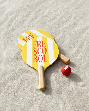 Two yellow and white striped beach paddles and a red ball lie on sand, ready for Frescobol play.