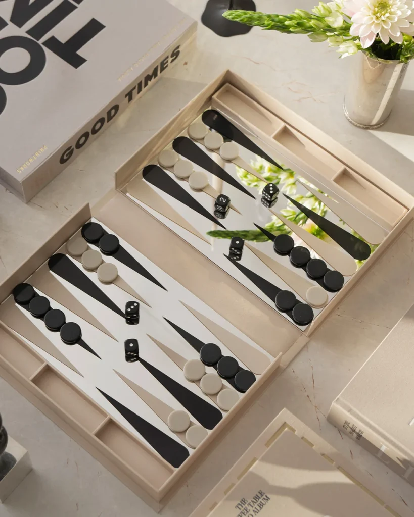 Open backgammon set with black and white checkers and dice on a light marble surface, with a flower vase nearby.