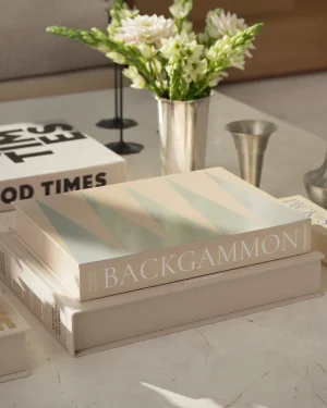 Stack of oversized neutral-toned books on a marble table, top book titled BACKGAMMON with a pastel striped cover; white flowers in a metal vase in the background.