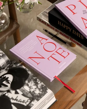Pink hardcover book titled 'A ONE' sits on a glass table beside a black 'Happy Days' book and a pen. A red bookmark ribbon protrudes from the pink book. Flowers in glass vases are in the background.