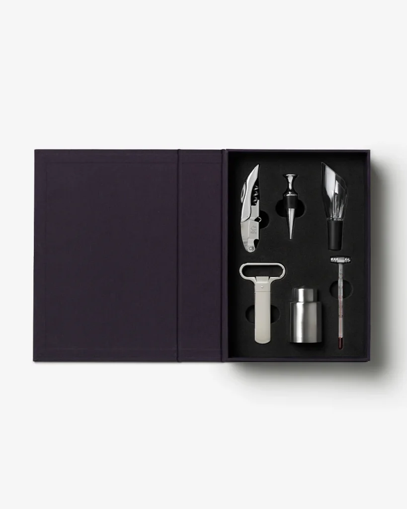 Open dark purple gift box revealing a stainless steel wine accessory set in foam: corkscrew/lever, foil cutter, pourer, stopper, keeper/vent, and a thermometer.