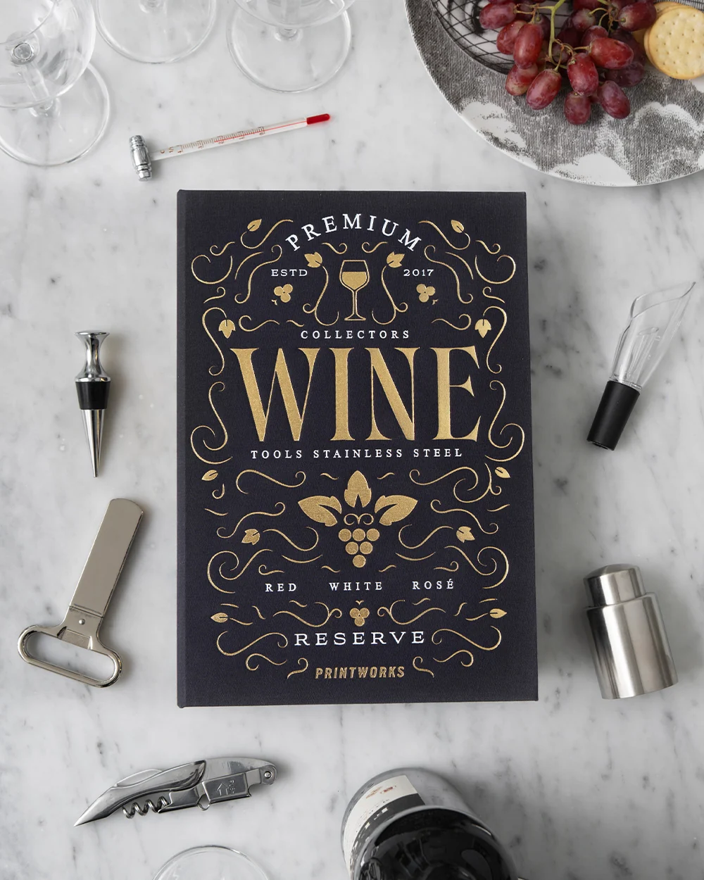 IMG_2533 - New Trend Dark blue wine book with gold lettering on a marble table, surrounded by wine accessories and a grape plate in the corner.