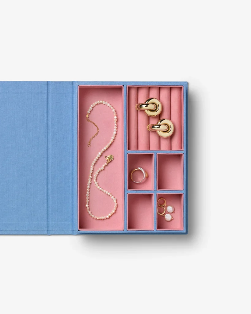 Open blue jewelry box with pink interior compartments displaying a pearl necklace, plus gold rings, earrings, a bracelet, and cufflinks neatly arranged.