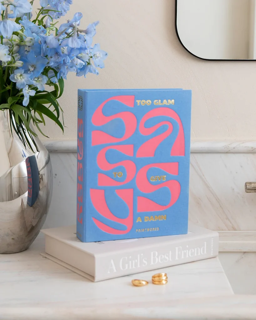 Blue decorative book with pink lettering 'Too Glam to Give a Damn' resting on a white book on marble, beside a vase of blue flowers.