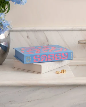 Two books stacked on a marble surface with a blue cover reading 'Sassy'; gold rings lie nearby and a vase of blue flowers sits to the left.