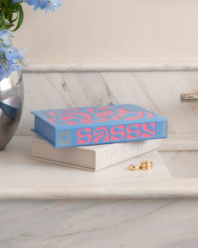 Two books stacked on a marble surface with a blue cover reading 'Sassy'; gold rings lie nearby and a vase of blue flowers sits to the left.