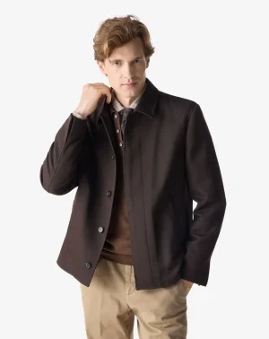 Male model wearing a dark brown coat over a brown sweater and tan chinos, posing with hand near his collar