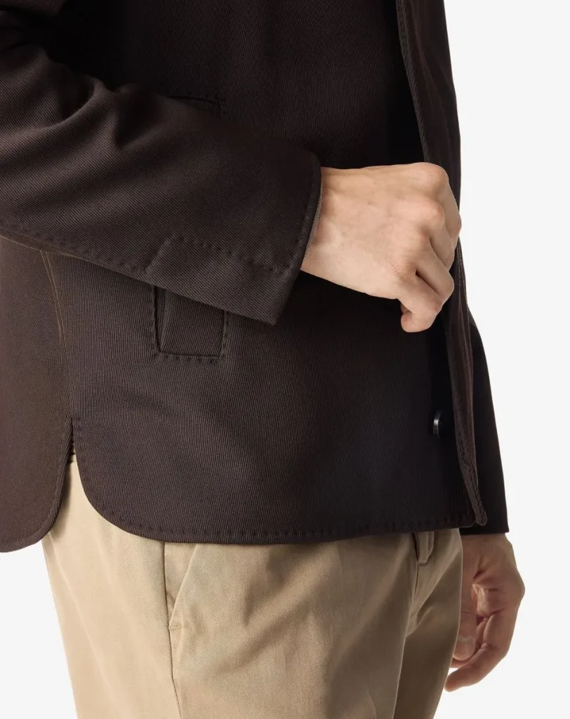 Close-up of a person wearing a dark brown blazer with a hand resting in the blazer pocket, beige pants visible below.