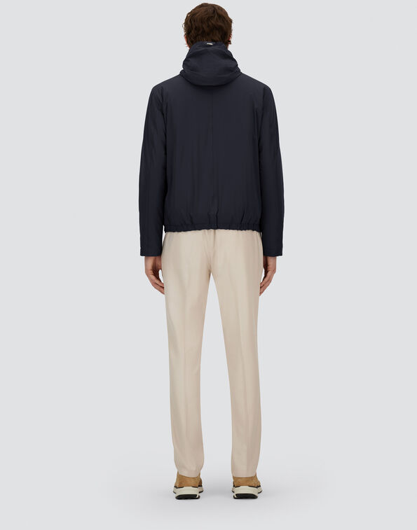 Back view of a person in a navy hooded jacket and beige trousers on a light gray backdrop.