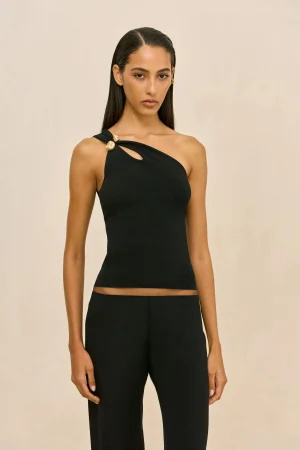 Woman posing in a black one-shoulder top with gold button accents against a beige backdrop, minimal jewelry visible.