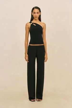 Full-body portrait of a woman in a black one-shoulder top with gold button accents and matching wide-leg pants, standing against a beige backdrop with sandals visible.