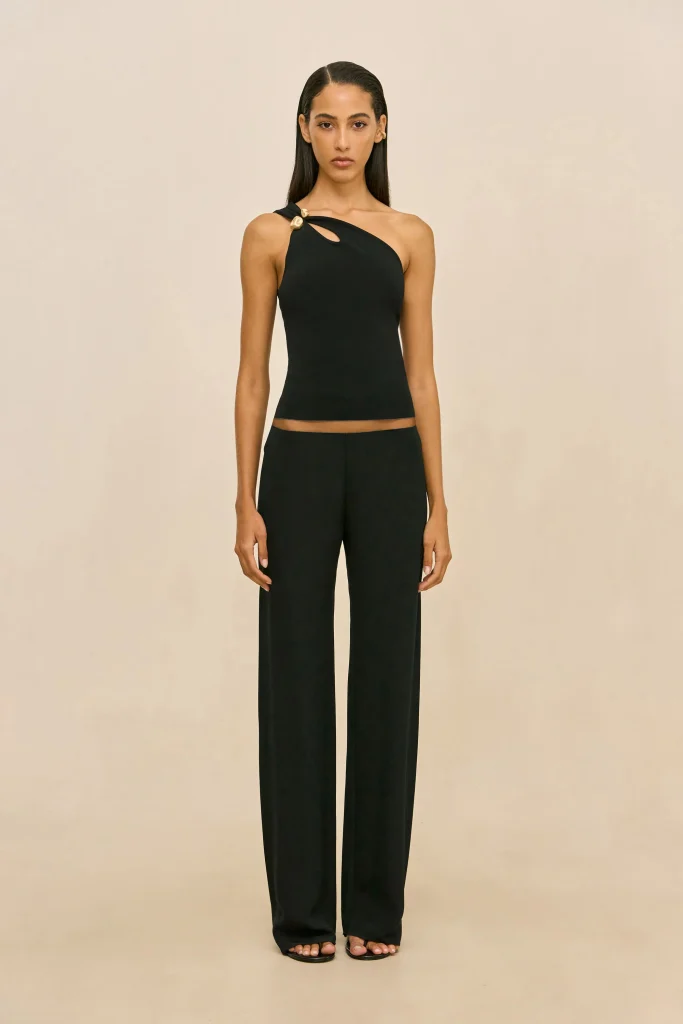 Full-body portrait of a woman in a black one-shoulder top with gold button accents and matching wide-leg pants, standing against a beige backdrop with sandals visible.