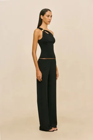 Woman in a fitted black sleeveless top with a cutout detail and matching wide-leg pants, standing against a beige backdrop, in profile view.