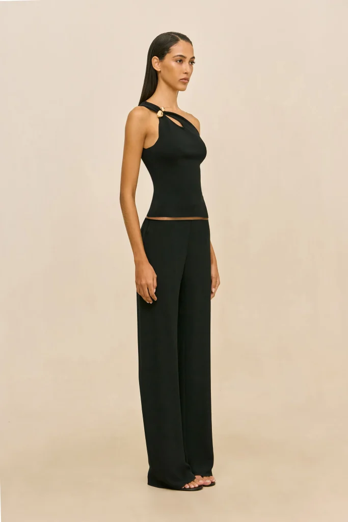 Woman in a fitted black sleeveless top with a cutout detail and matching wide-leg pants, standing against a beige backdrop, in profile view.