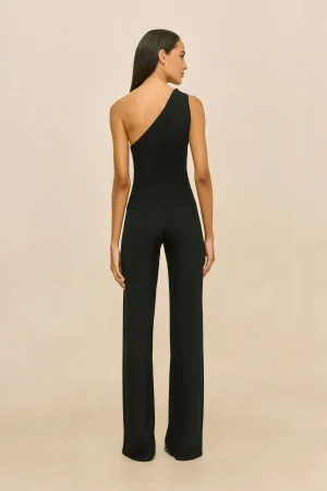Woman standing with her back to the camera, wearing a black one-shoulder sleeveless top and wide-leg black pants against a beige backdrop.