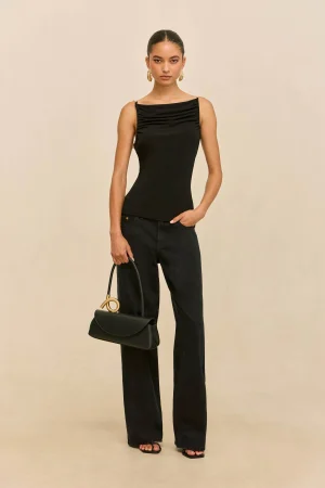 Full-body portrait of a woman wearing a black sleeveless top with ruching at the neckline, black wide-leg pants, gold hoop earrings, and a black handbag with a gold circular handle against a beige background.
