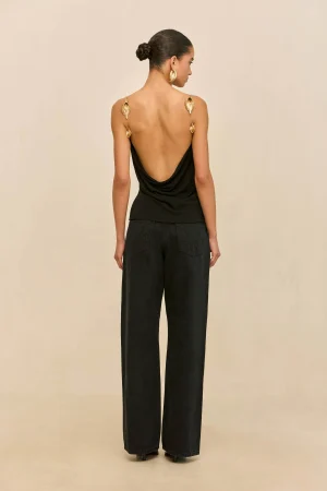 Back view of a woman wearing a black draped open-back top, gold leaf earrings, and wide-leg black pants.